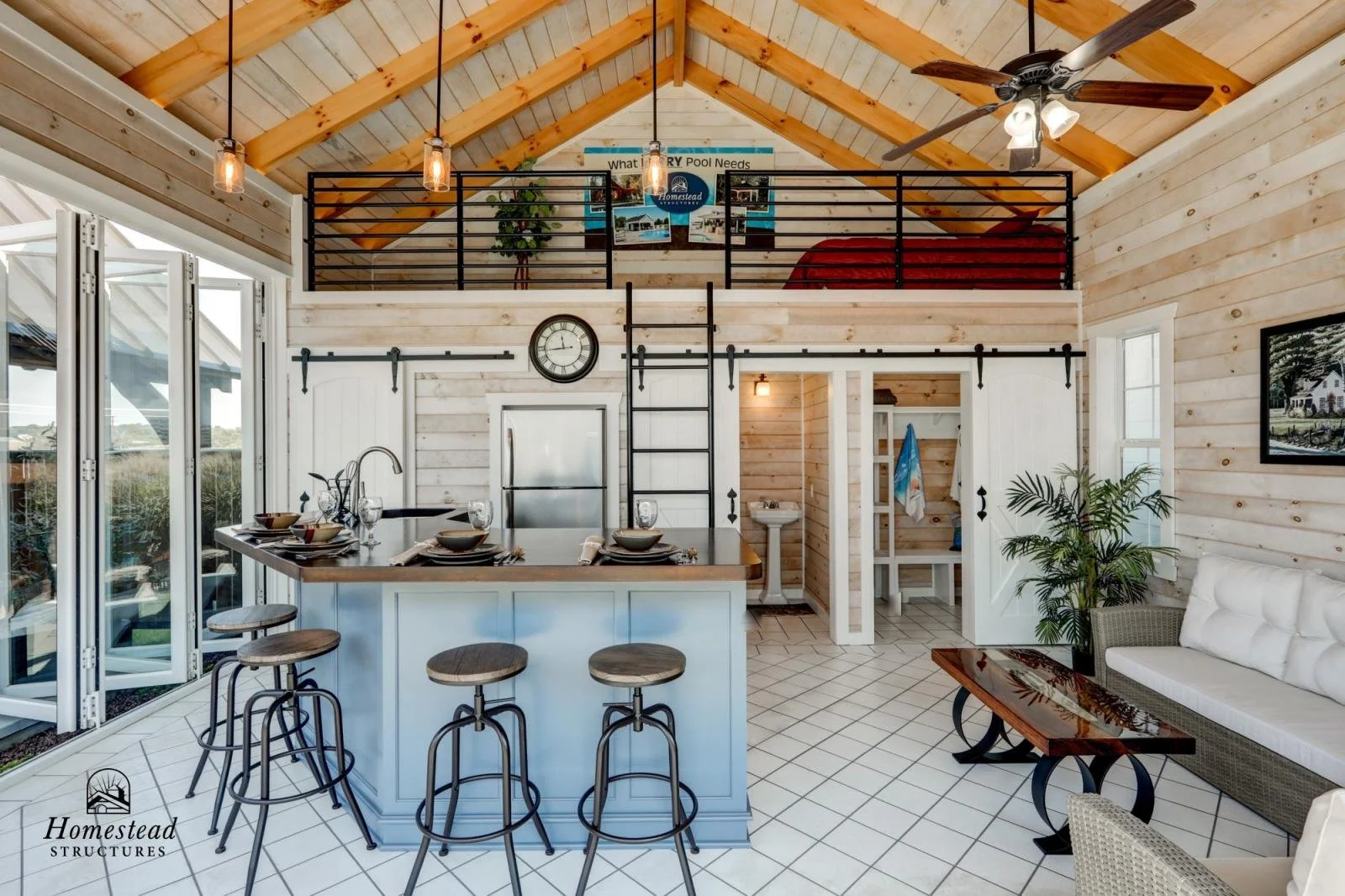 Interior view of a cozy, rustic-style living space with a high, sloped wooden ceiling, ceiling fan, and pendant lights. There is a kitchen with a small island and four bar stools, a white refrigerator, and sliding glass doors. An open loft with a rai