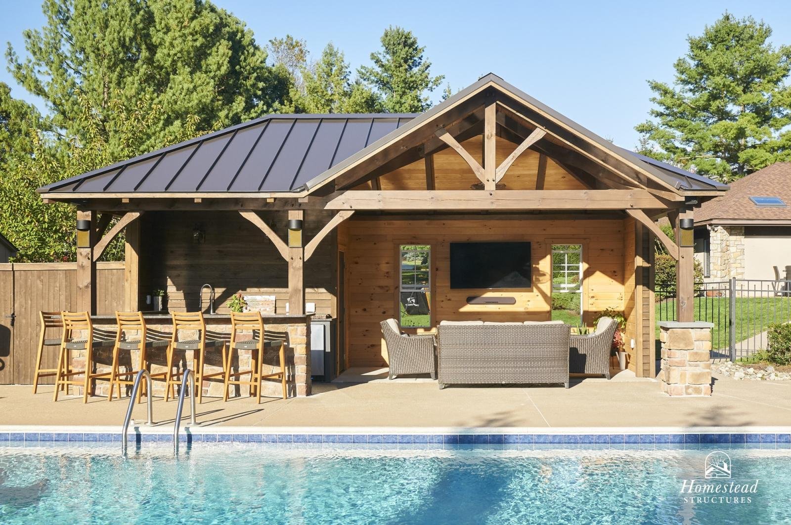 Outdoor pool area with a built-in bar, and a wooden cabana with seating and a wall-mounted TV, surrounded by trees and neighboring houses.
