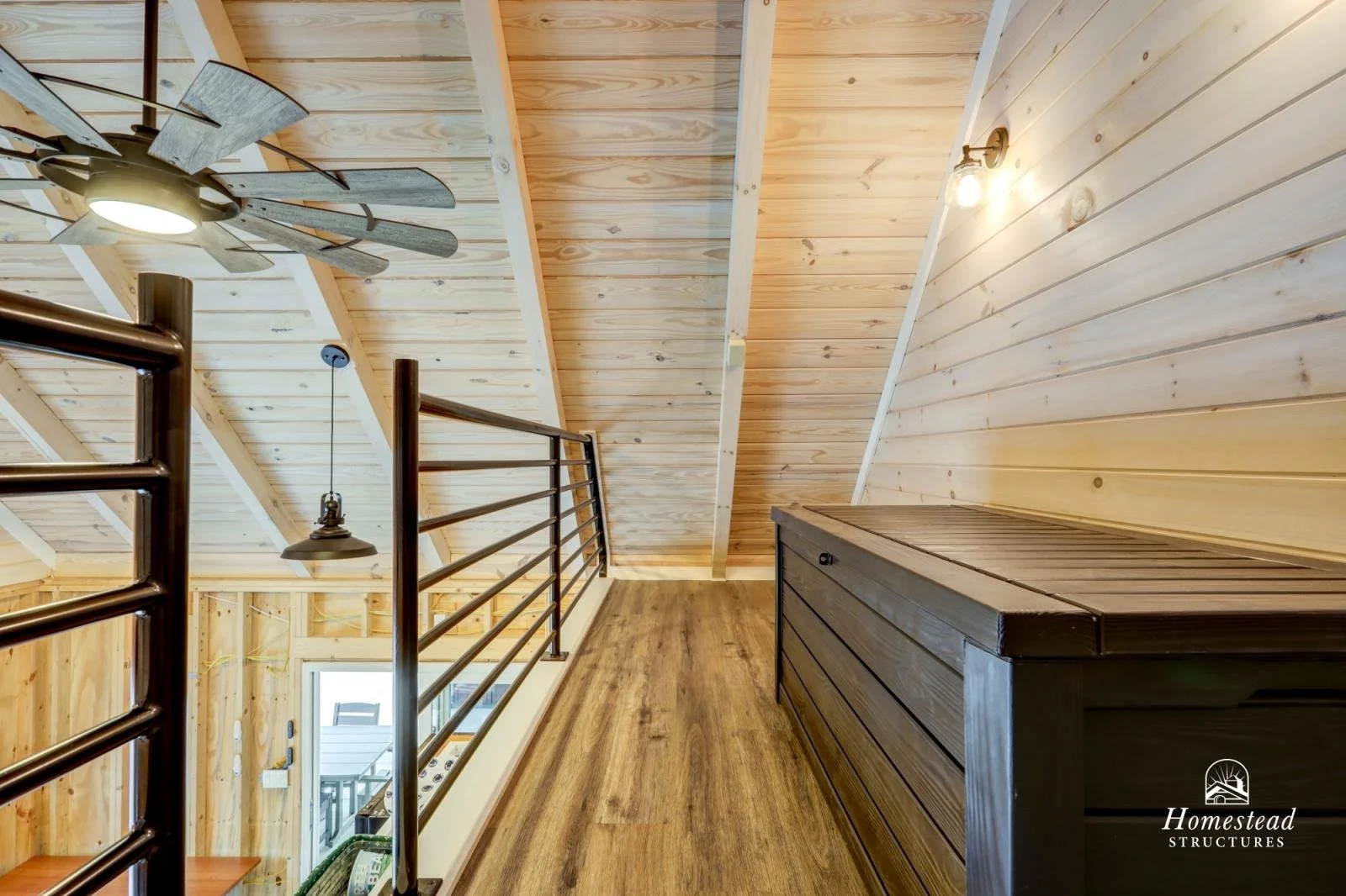 Wooden interior loft with sloped ceiling, ceiling fan, hanging light, black railing, and wooden storage chest