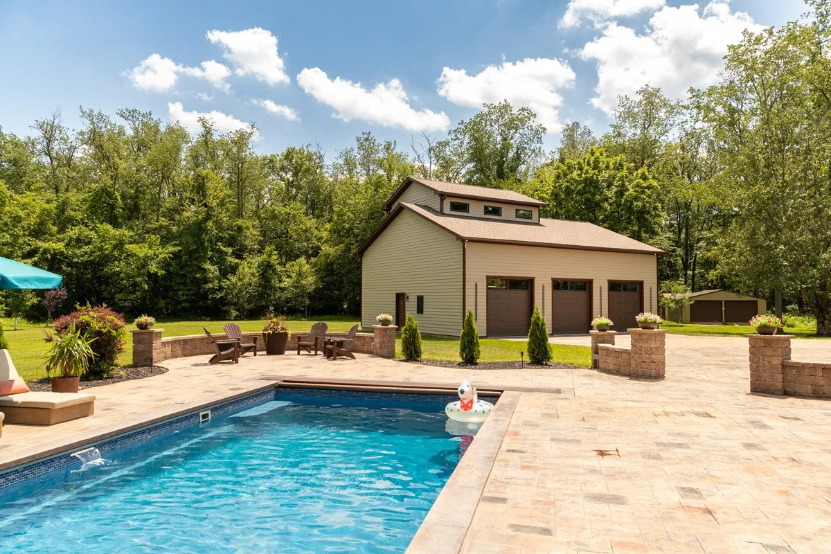 Residential backyard with swimming pool, Adirondack chairs, potted plants, a tan garage with three doors, and a treeline in the background under partly cloudy skies.