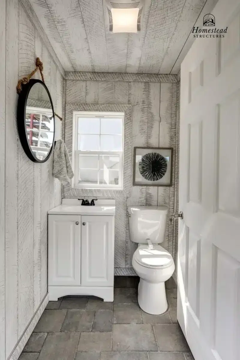 A small bathroom with wood-textured walls and ceiling, a white vanity with black faucet, a toilet, a window, a round mirror hanging on a rope, a framed artwork of a black spherical design, and gray tiled flooring.