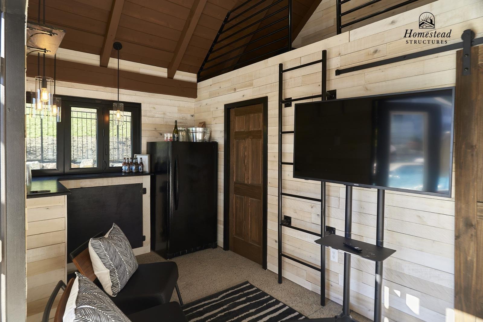 Interior of a modern, rustic living space with a black refrigerator, a flat-screen TV on a metal stand, a wooden door, and a window with black framing, featuring wood-paneled walls and a sloped wooden ceiling.