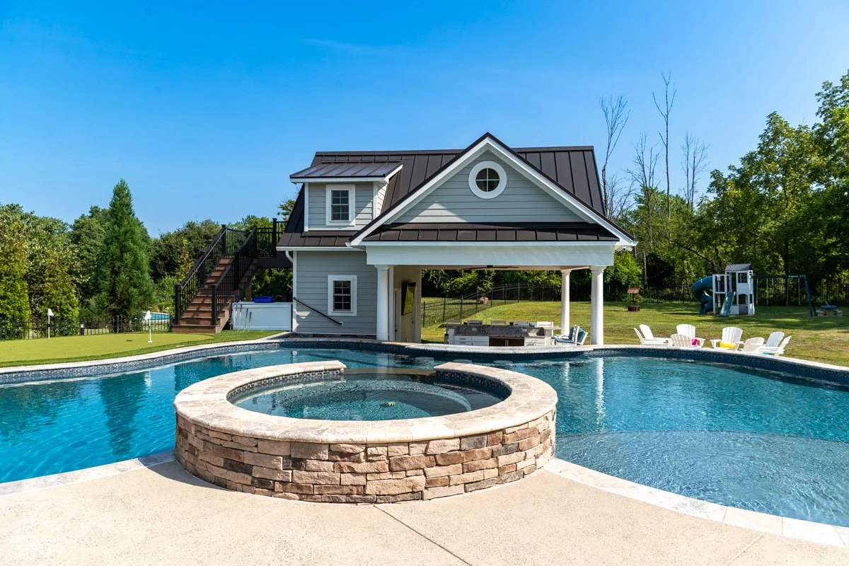 Backyard with swimming pool, hot tub, and a pool house with stairs leading to an upstairs deck, surrounded by green trees and lawn, with outdoor chairs and a playground in the background.