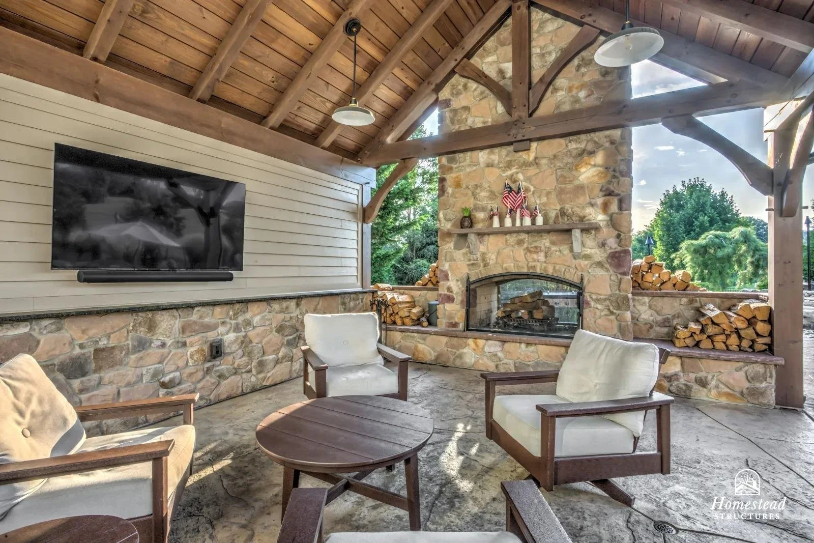 Outdoor living area with a stone fireplace, wood logs stacked on the sides, a mounted flat-screen TV, and four wooden armchairs with cream cushions around a round wooden table, under a wooden ceiling with pendant lights. Decor on the fireplace mantle
