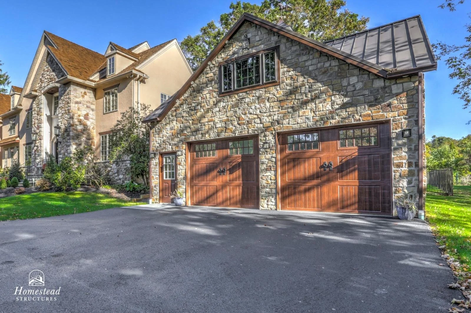A multi-story house with stone and stucco exterior, steep gabled roof, and wooden garage doors, surrounded by green lawn and trees, under a clear blue sky.