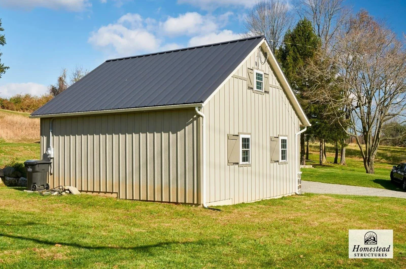 A beige metal-sided barn with a dark gray metal roof, set in a grassy field with trees in the background, and a gravel driveway to the side.