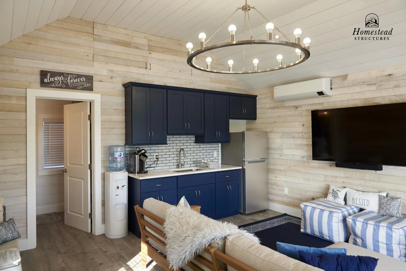 Interior of a cozy living room with a blue kitchen cabinet, a small sink, a refrigerator, a water dispenser, a wall-mounted TV, and a sitting area with cushions. A chandelier hangs from the ceiling, and the walls are made of light-colored wood planks