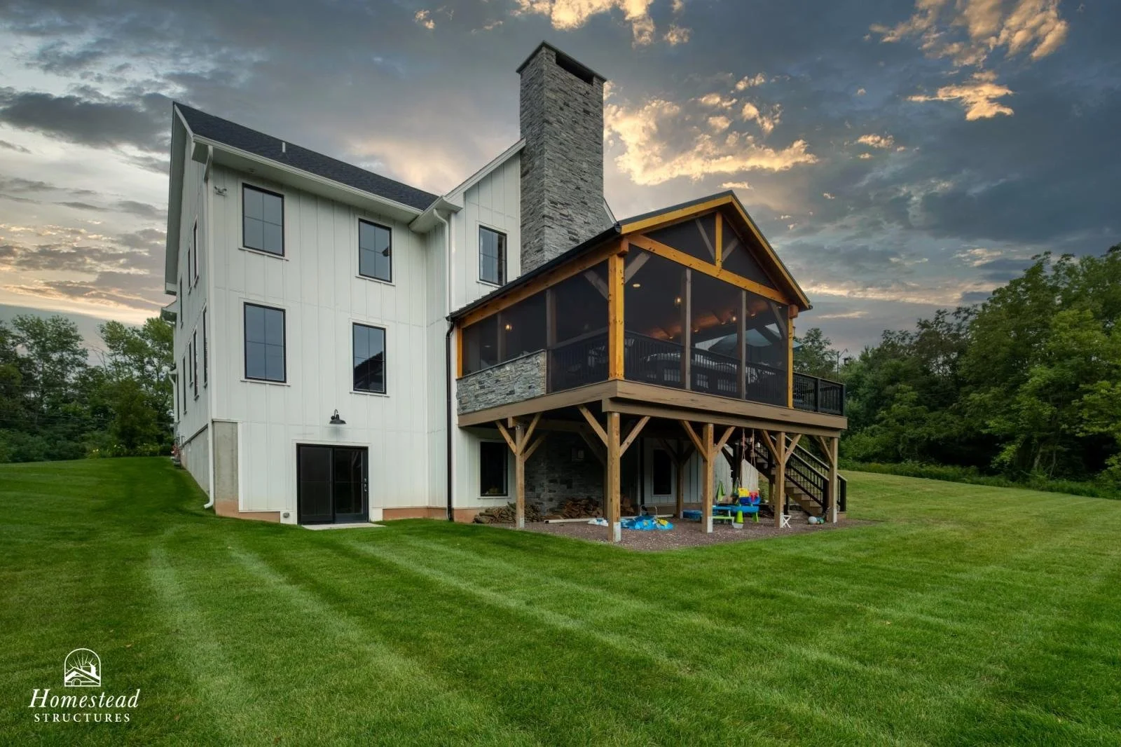 A large white modern house with multiple windows and a black door, featuring a screened-in porch with wooden beams and a stone chimney, set on a well-manicured lawn surrounded by trees at sunset.