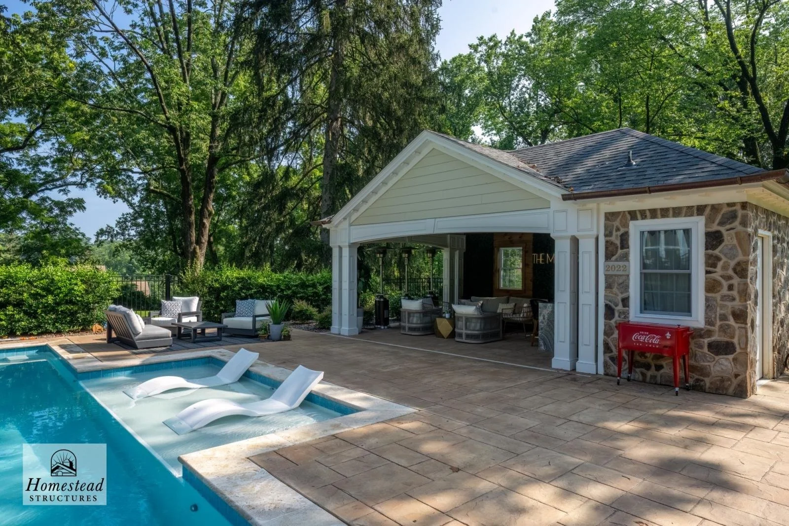 Outdoor backyard area with swimming pool, lounge chairs, seating area with cushioned chairs, and a small building with stone walls, white trim, and a Coca-Cola cooler outside. Surrounded by green trees.