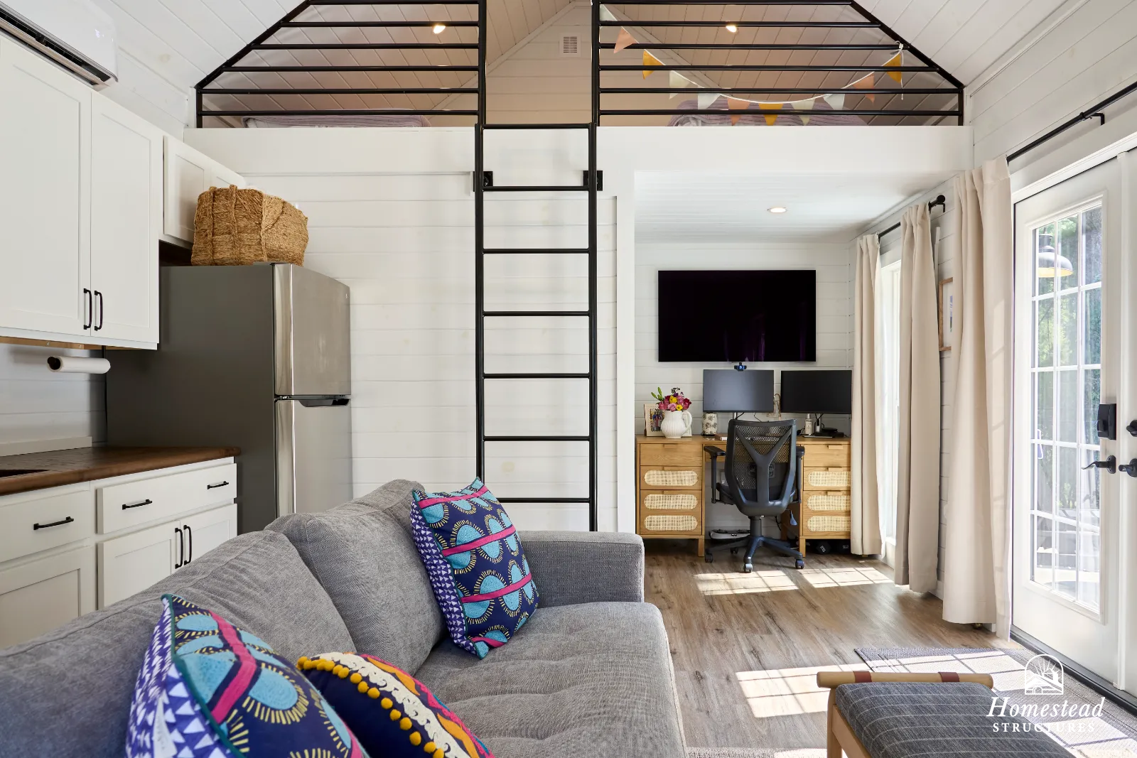 Living room with a gray couch with colorful pillows, a wooden desk with two computer monitors, a black office chair, and a large flat-screen TV mounted on a white wall with curtains on the window and glass door leading outside.