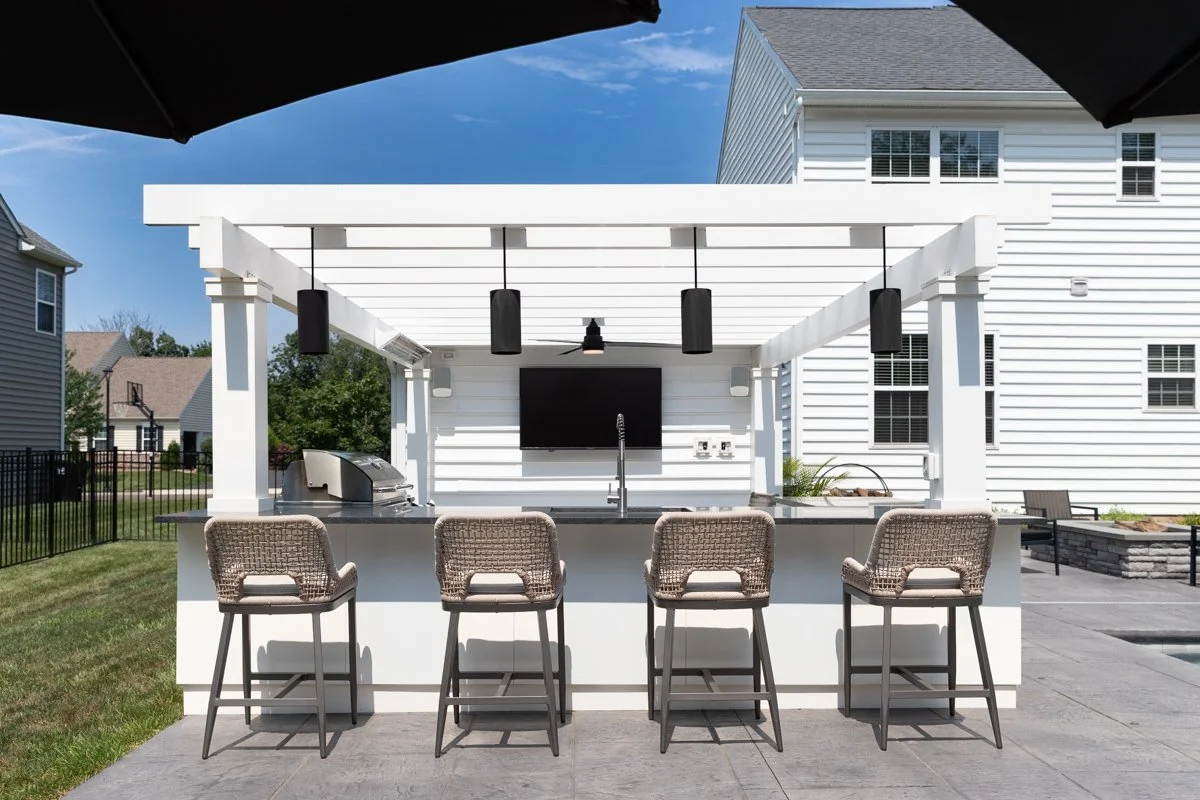 Outdoor kitchen area with four barstools, a TV, and a grill under a white pergola, in a residential backyard