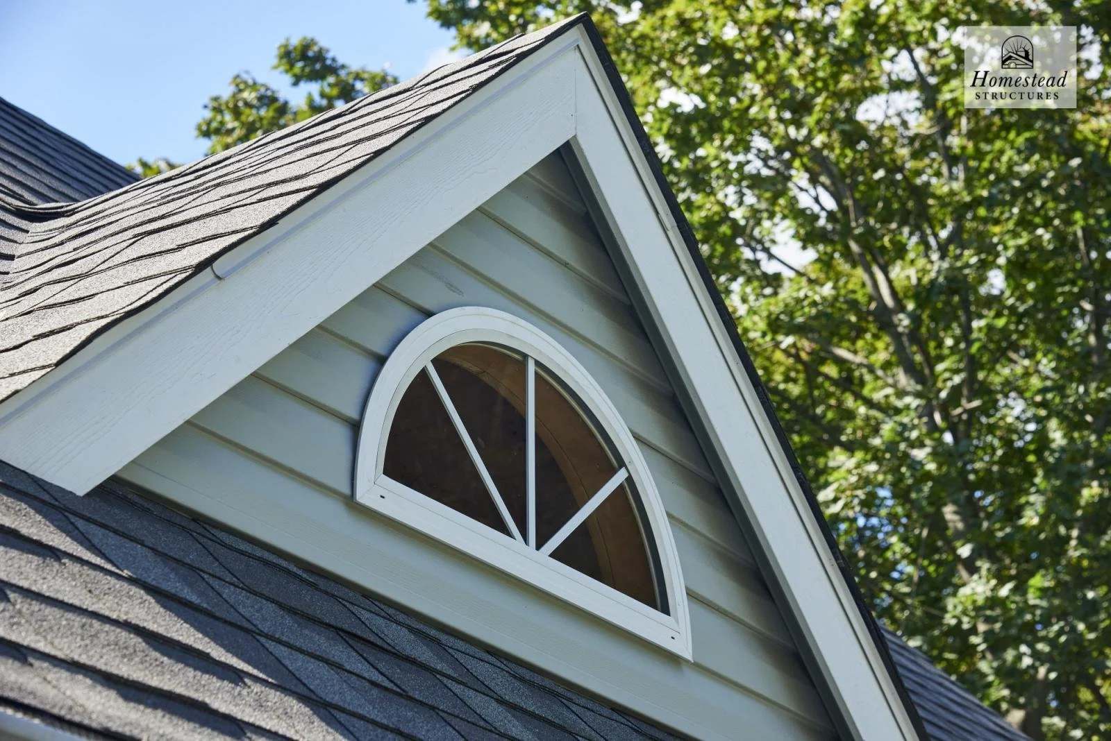 Close-up view of a house's gable with a semi-circular window, beige siding, gray shingles on the roof, and trees in the background.