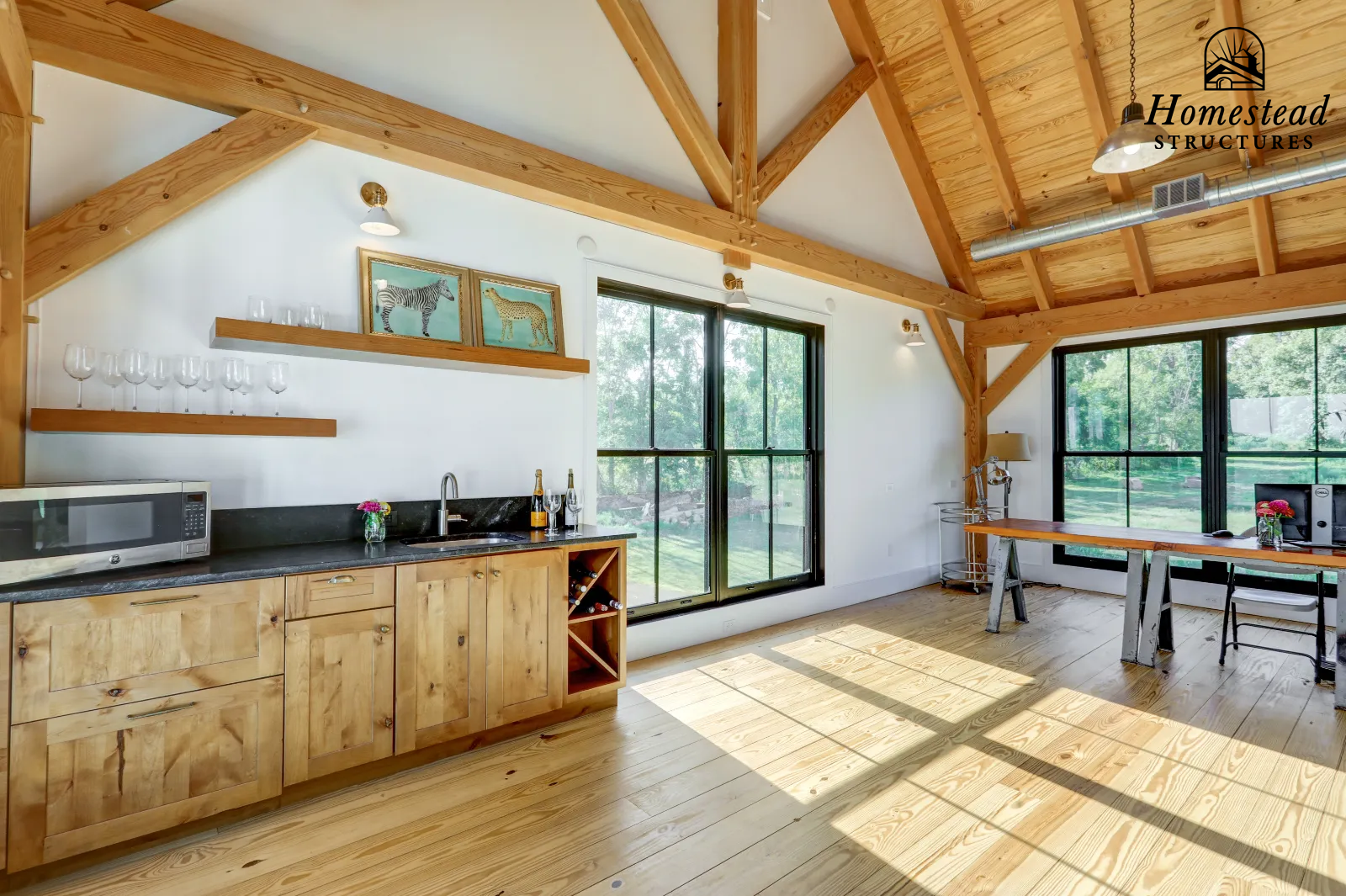 Open kitchen and dining area with wooden beams and flooring, large windows letting in sunlight, a kitchen counter with a microwave, wine bottles, glasses, and a small flower arrangement; wall shelves with glasses and artwork of zebras and cheetahs; t