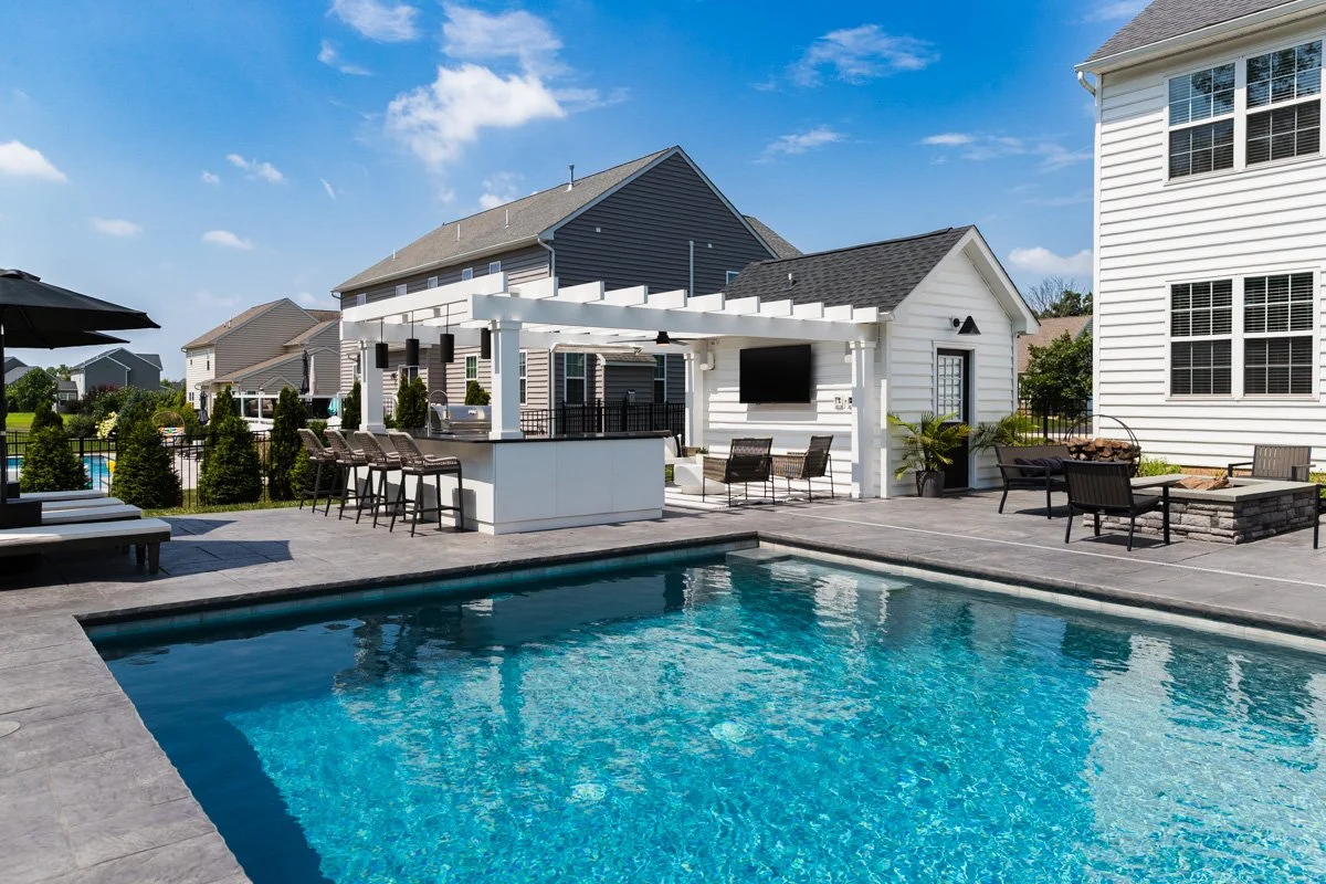 Residential backyard with a swimming pool, patio furniture, a bar area with barstools, and a poolside outdoor TV under a pergola.