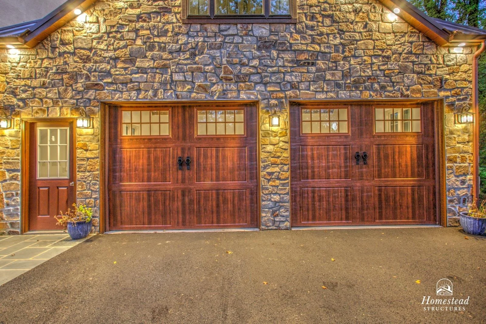 A stone and wood garage door with two small windows at the top, to the right of a small wooden door with a window, on a house's exterior wall illuminated by wall-mounted lights. Two potted plants are near the doors.