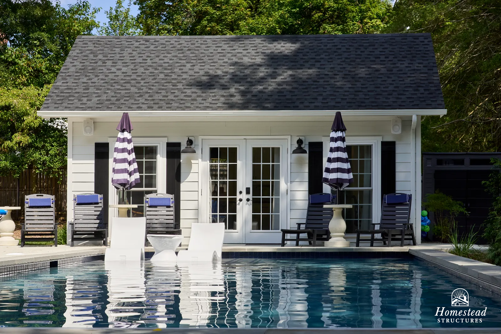 A backyard scene featuring a swimming pool in the foreground, a small white house with black shutters and a glass double door in the background, surrounded by trees, with outdoor chairs and umbrellas on the patio.