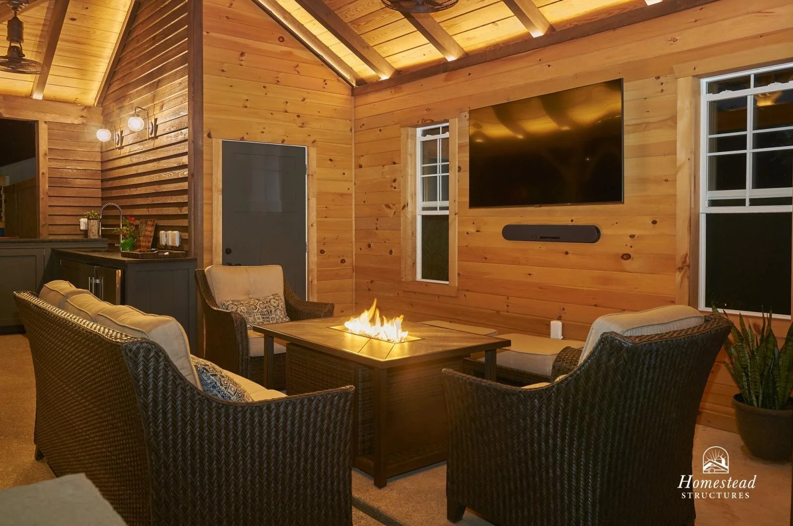 Living room with wooden walls and ceiling, furnished with wicker chairs and a central fire feature, a large wall-mounted TV with a speaker beneath it, and windows with night outside view. There is a kitchenette or bar area on the left with a sink and