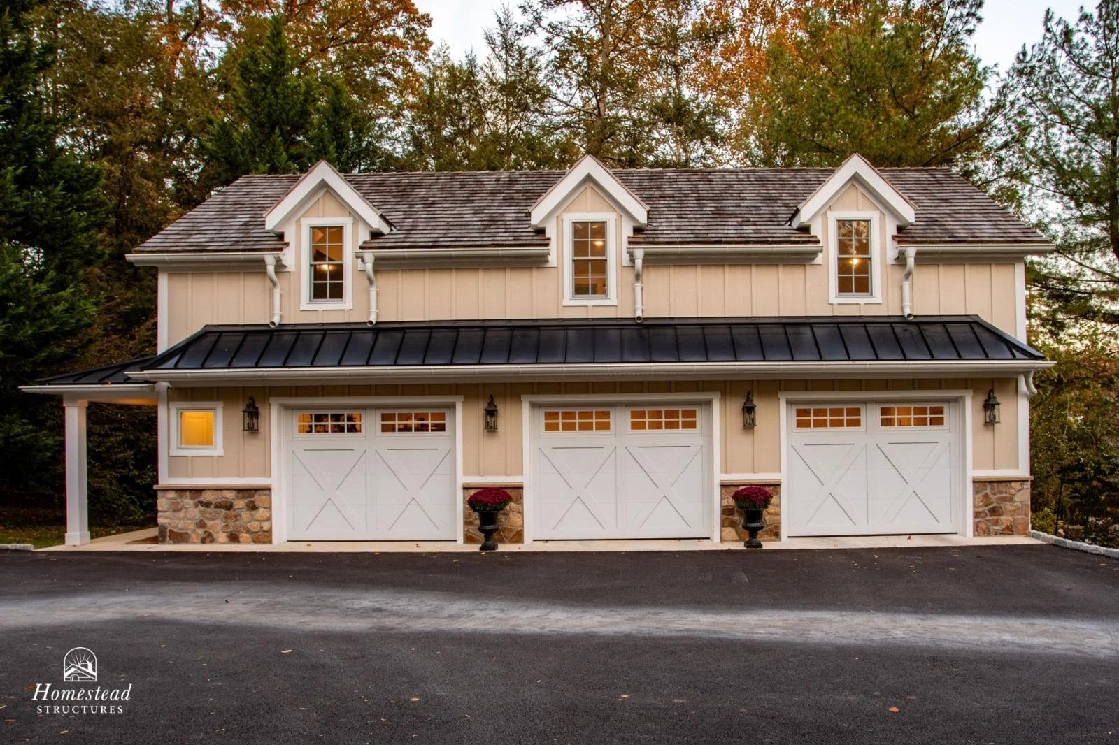 A beige two-story garage with three white paneled garage doors, stone accents at the base, black metal roof, and three small dormer windows on the upper level, surrounded by trees.