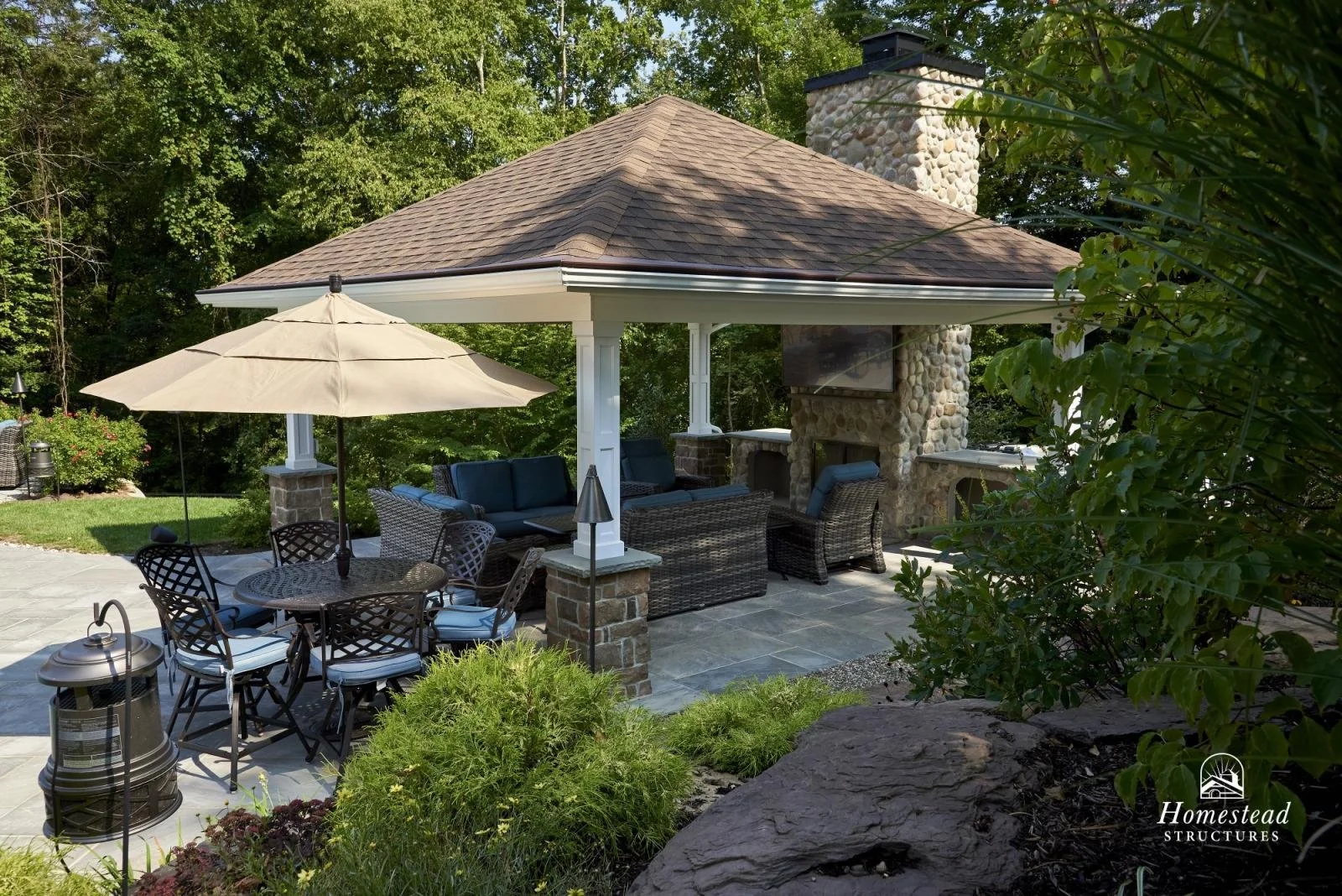 Outdoor patio area with wicker furniture, a large umbrella, a stone fireplace, and a television under a gazebo-style roof surrounded by greenery.