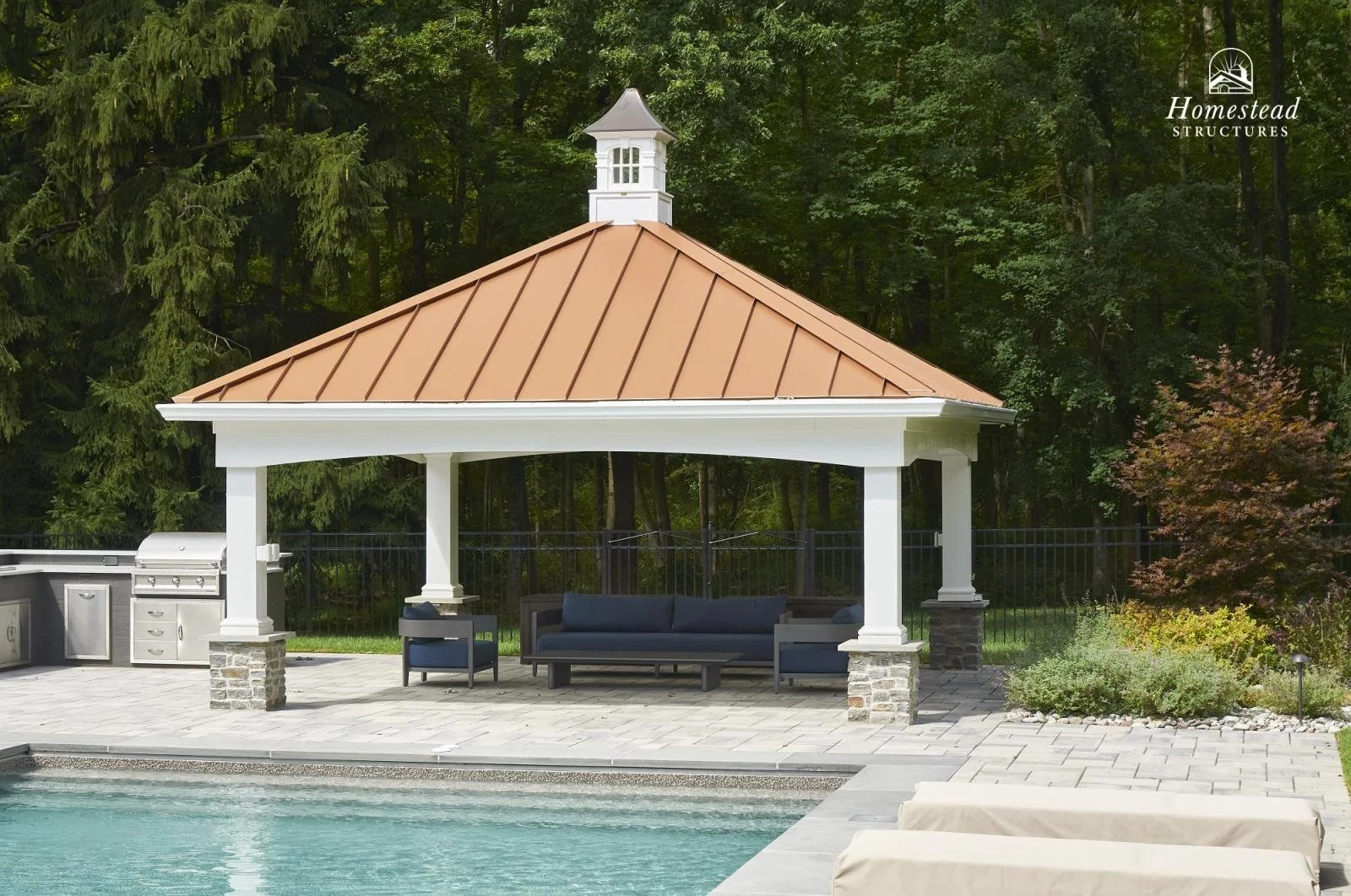 Outdoor pool area with a beige umbrella-covered seating area, barbecues, and lush green trees in the background, surrounded by a black fence.