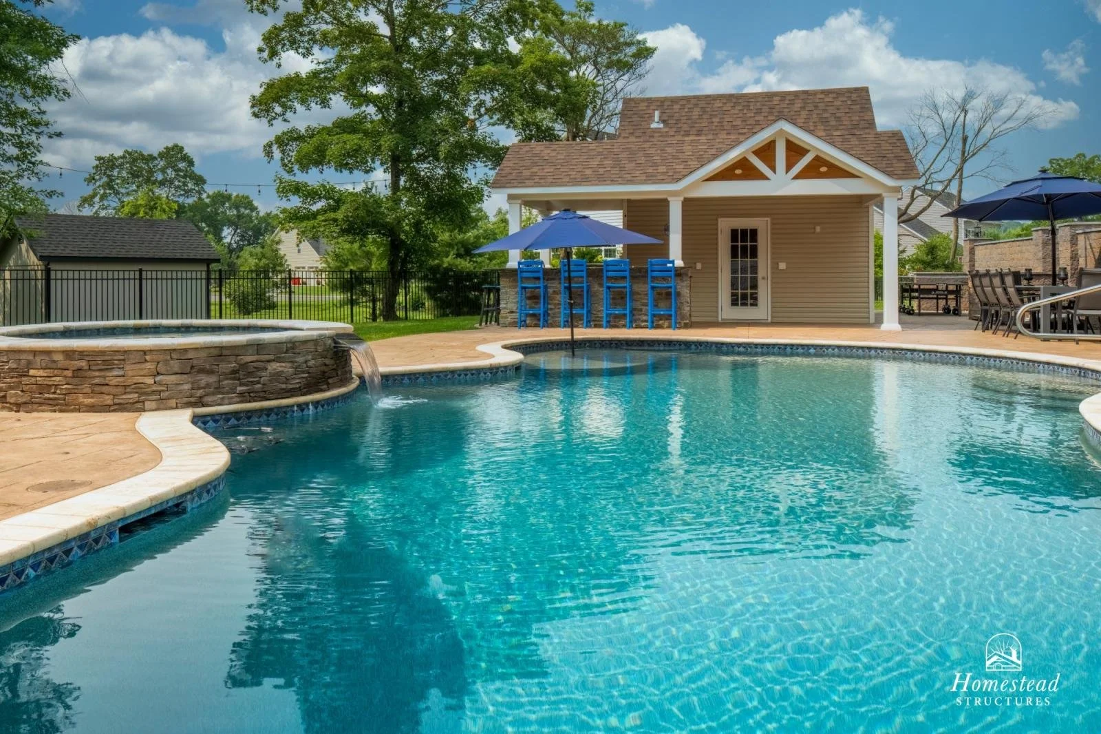 A backyard pool area featuring a large, curved swimming pool with a small waterfall fountain on the left, stone accents, and a deck with lounge chairs, umbrellas, and a small building with a bar area, surrounded by trees and grass under a partly clou