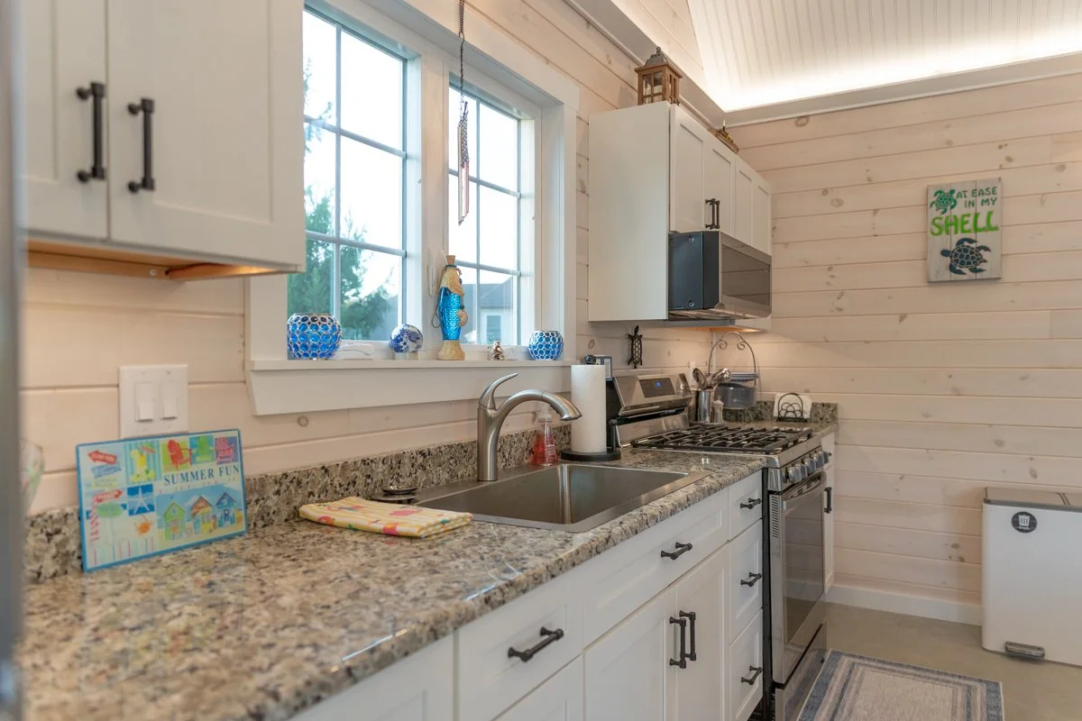 A cozy kitchen with white cabinets, granite countertops, and a window above the sink. Decorations include blue vases and a small lighthouse. A stove and microwave are on the right, with a wall art piece about a shell. The wall is paneled in light-col