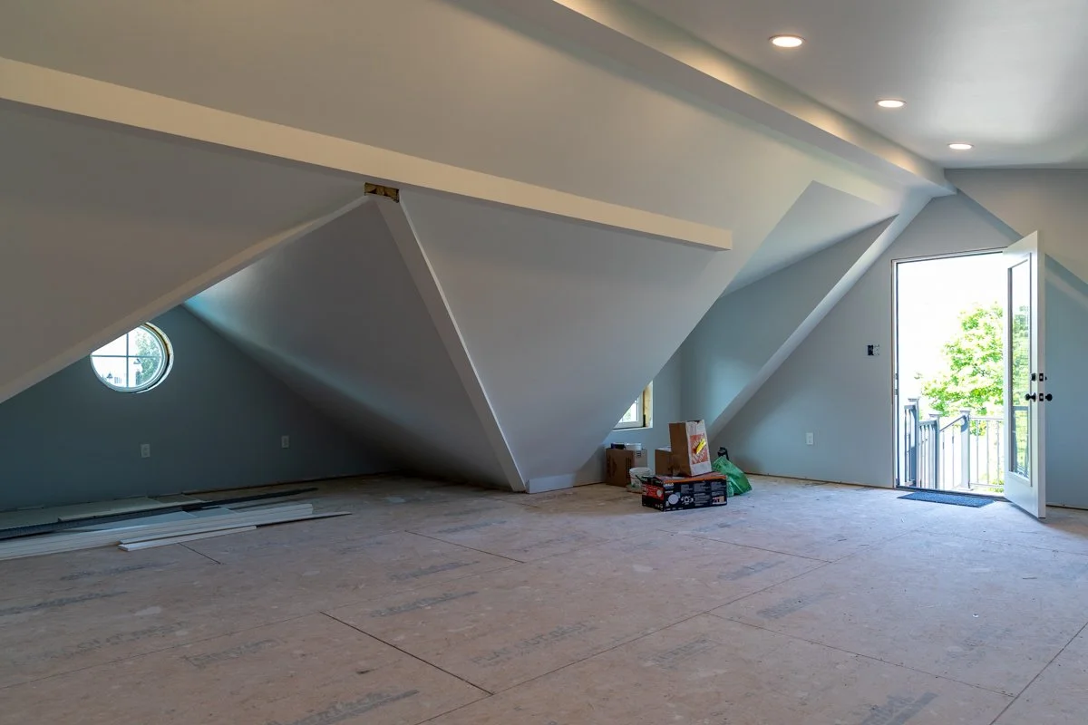 An attic or loft space under construction with sloped ceilings, a small circular window, a door leading outside, and unfinished flooring with construction materials and boxes.