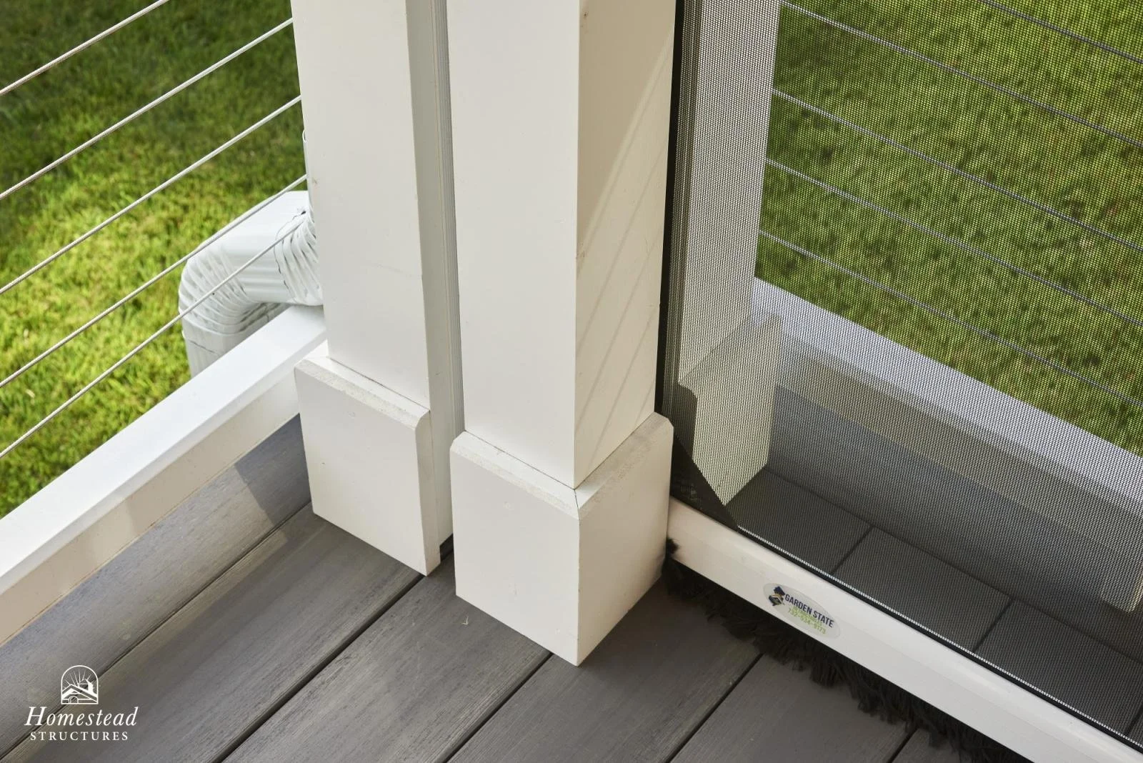 Close-up of a corner of a screened porch or deck, showing white wood posts and trim, a black mesh screen, and wooden deck flooring, with grass visible outside.
