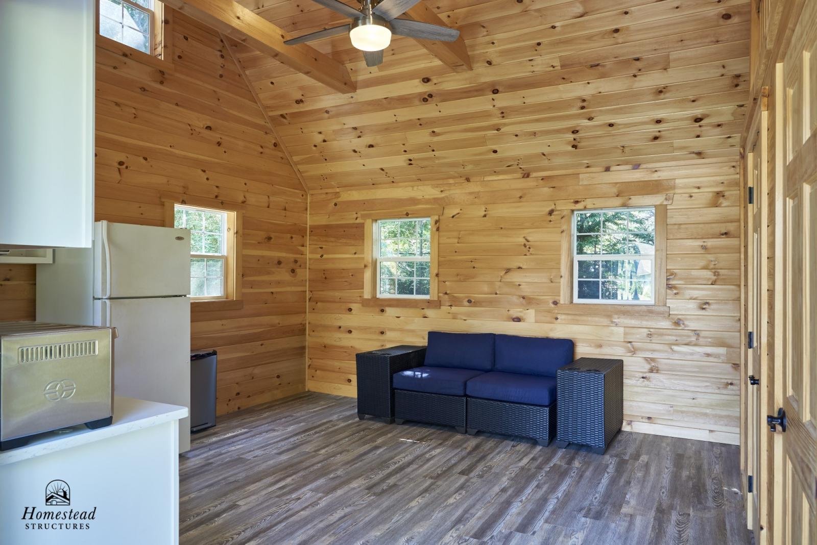 Interior of a wooden cabin with a black sofa, a mini fridge, and a toaster, with three small windows and a ceiling fan.