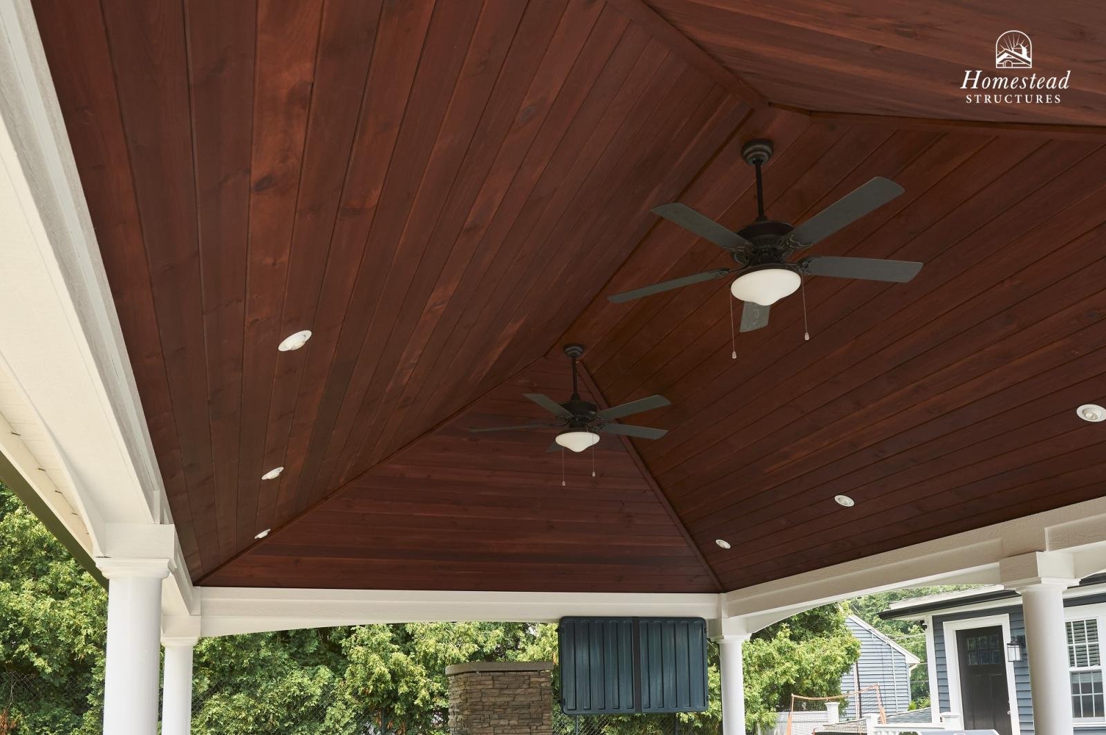 Covered porch area with a wooden ceiling, two ceiling fans, and white columns with a view of trees and neighboring houses in the background.