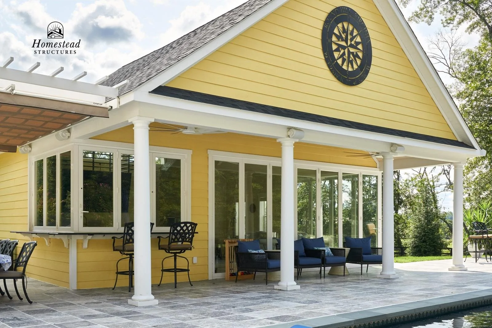 Yellow house with white columns, black clock on the gable, and a large screened porch with outdoor furniture.