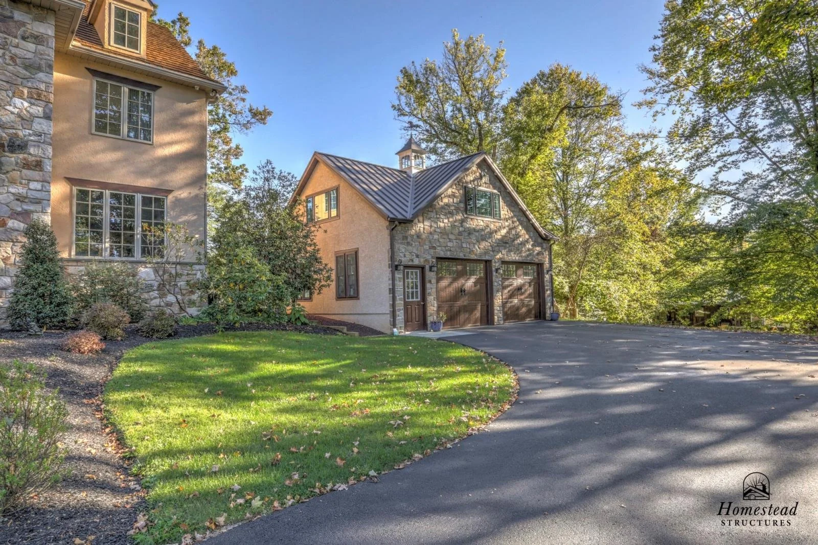 A house with beige stucco and stone exterior, a metal roof, and a driveway surrounded by green lawn and trees.
