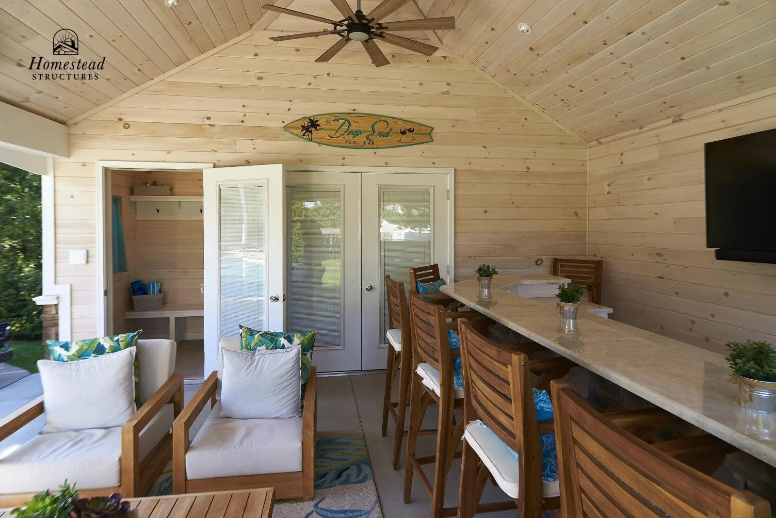 Indoor bar area with wooden panel walls and ceiling, bar counter with bar stools, wall-mounted TV, small seating area with white cushioned chairs, decorative pillows, potted plants, and a ceiling fan.