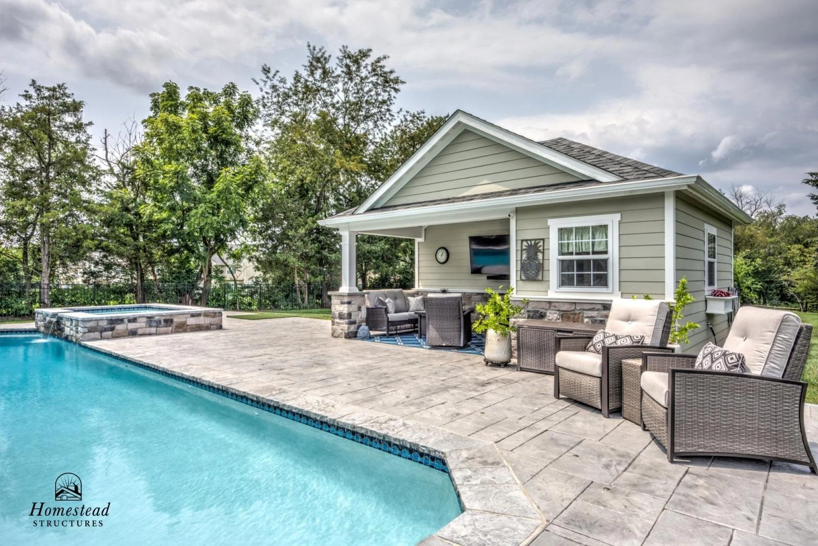 Backyard with a swimming pool, stone patio, outdoor seating, house, and trees in the background.