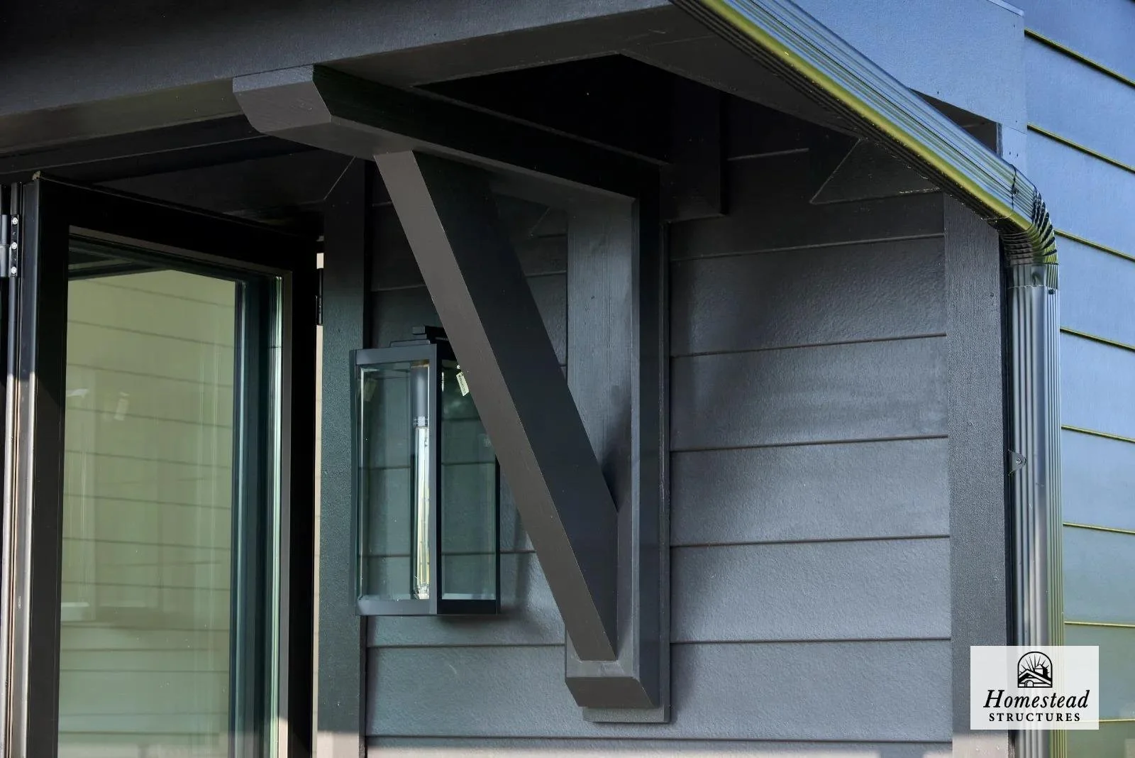 Close-up of a modern home exterior with black and gray siding, featuring a small black wall-mounted lantern, a window, and a gutter system.