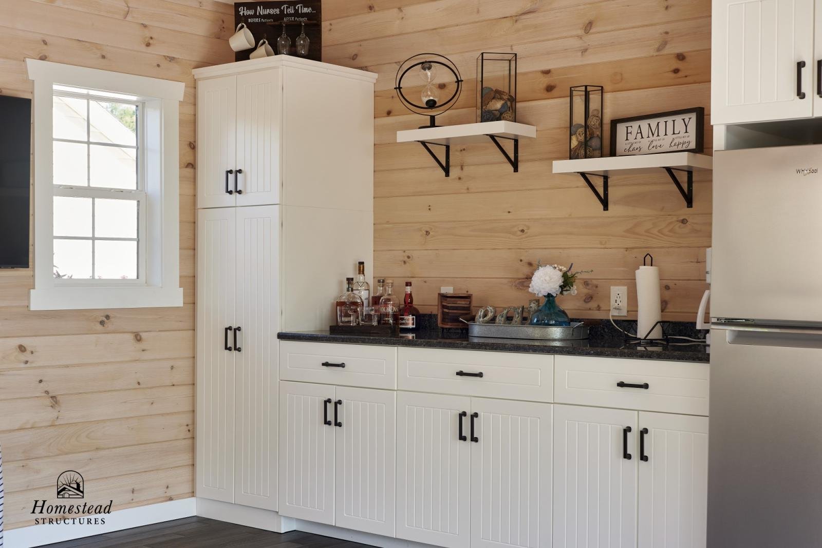 A rustic kitchen with light wood paneled walls, white cabinets, black countertop, window, and shelves with decorative items, including bottles, a play clock, and signs with text.