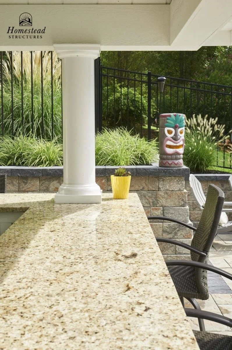 A patio with a granite counter, outdoor chairs, and a tiki statue, overlooking a garden with grasses and trees.