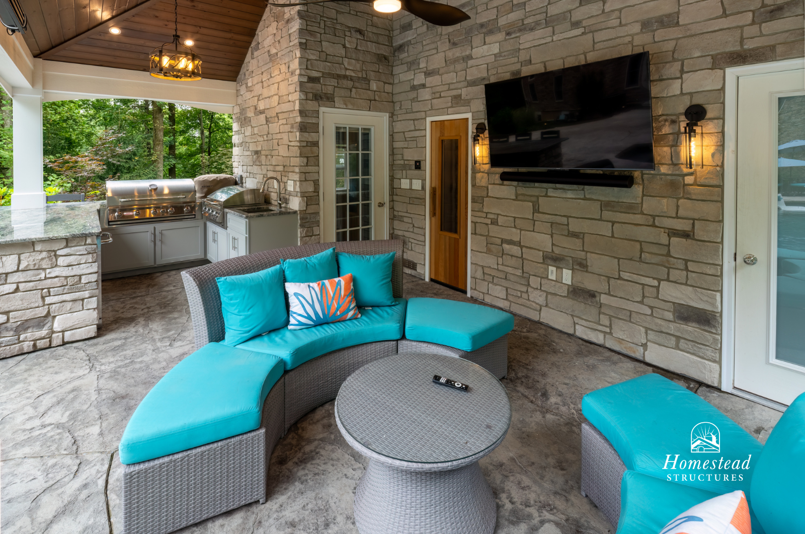 Outdoor living area with a curved sectional sofa with bright turquoise cushions, a round wicker table with a remote control, a wall-mounted TV, a stone wall and an outdoor kitchen with countertops and a grill, with lush greenery visible through the o