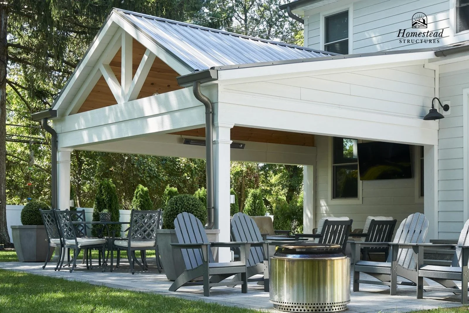 Backyard patio area with outdoor furniture in front of a house with siding and a TV, surrounded by trees and shrubs.