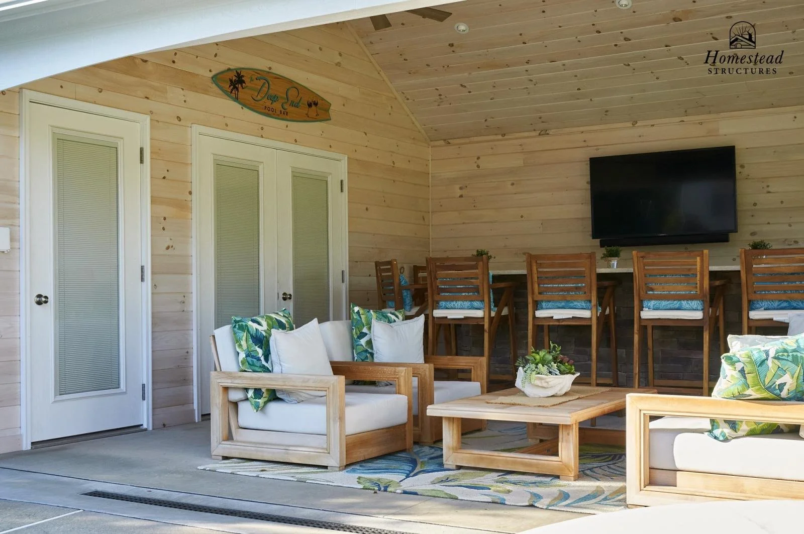 Indoor porch area with wooden furniture, tropical-themed cushions, potted plants, a large TV mounted on wooden-paneled wall, and a sign reading "Deep Sea Pool Bar".