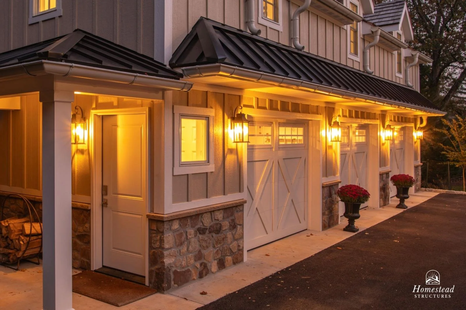 The exterior of a house with a garage door, illuminated by yellow exterior wall lights at dusk. The house has a wood and stone facade, and two large potted red plants are placed near the garage.
