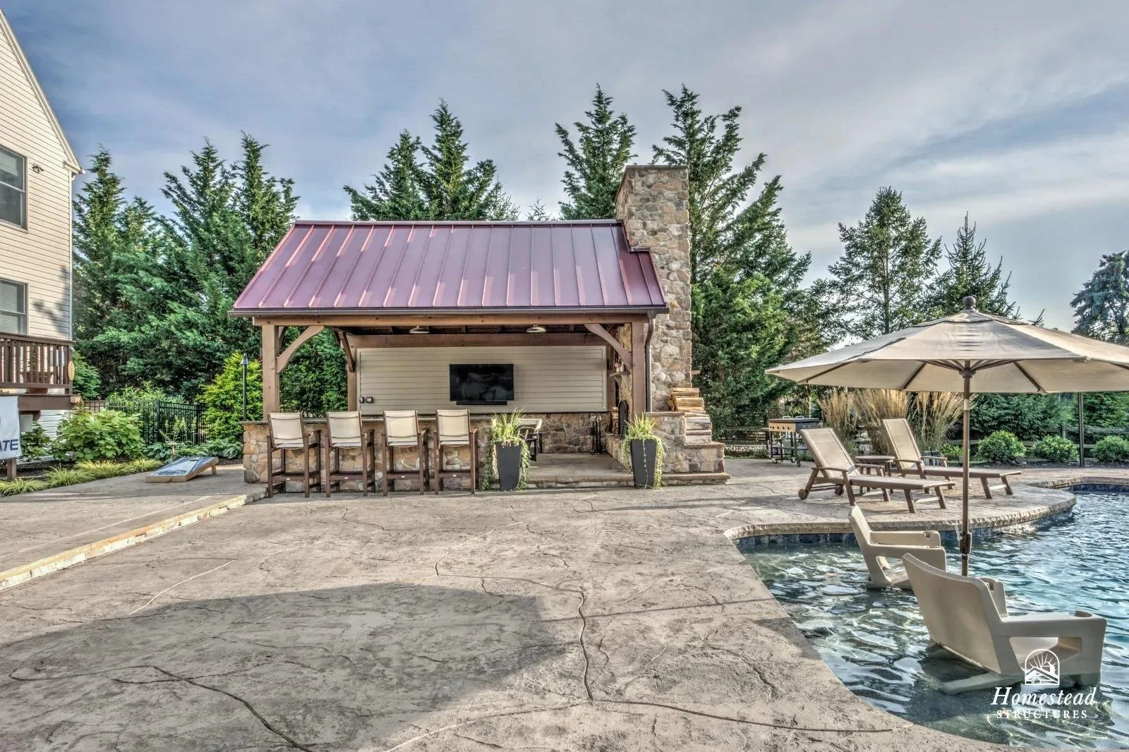 Outdoor backyard pool area with lounge chairs, a large umbrella, and a covered bar area with barstools, surrounded by greenery and tall trees.