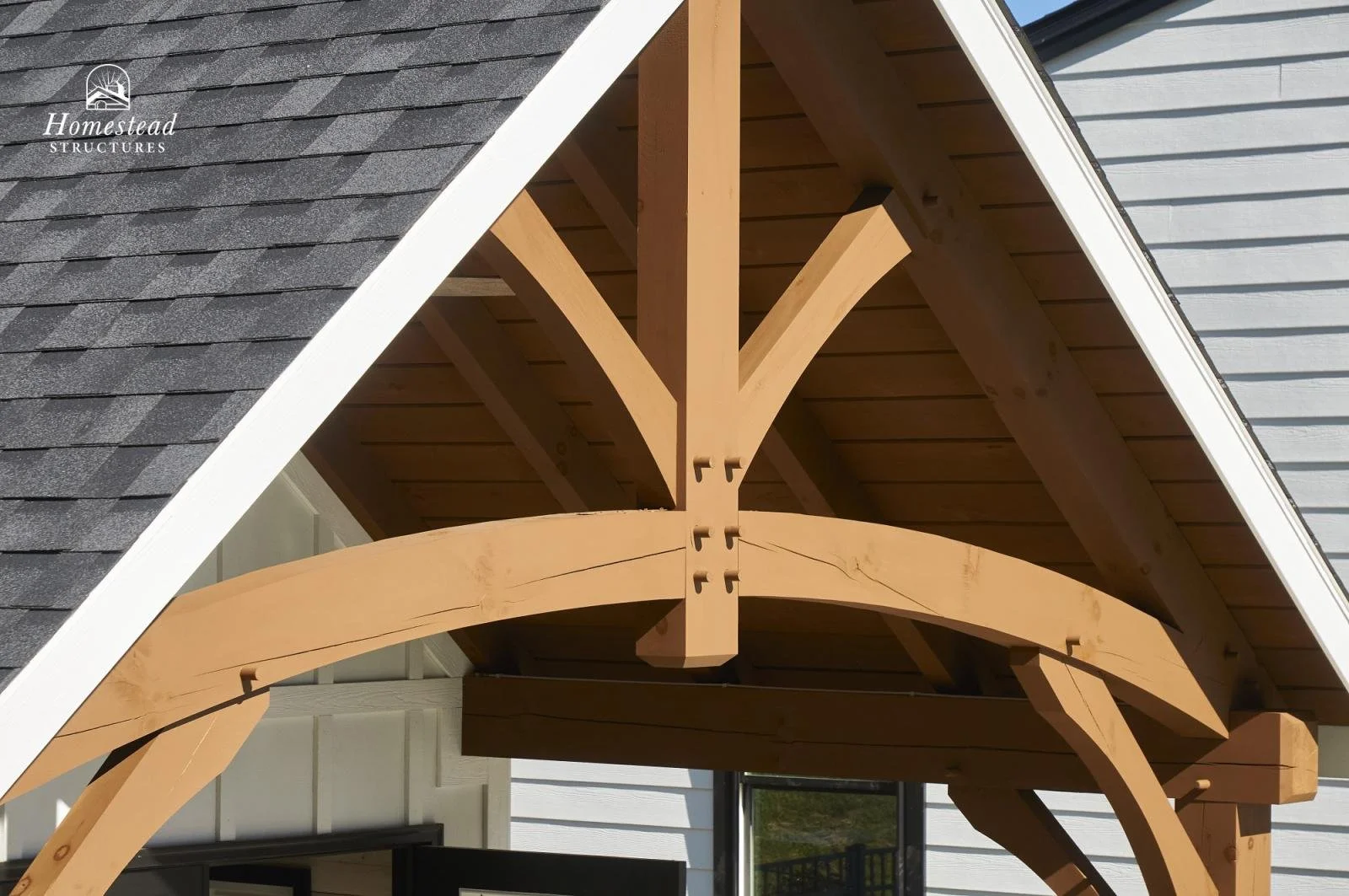 A close-up view of a wooden arch structure supporting a roof overhang, with shingles on the roof and a house wall in the background.