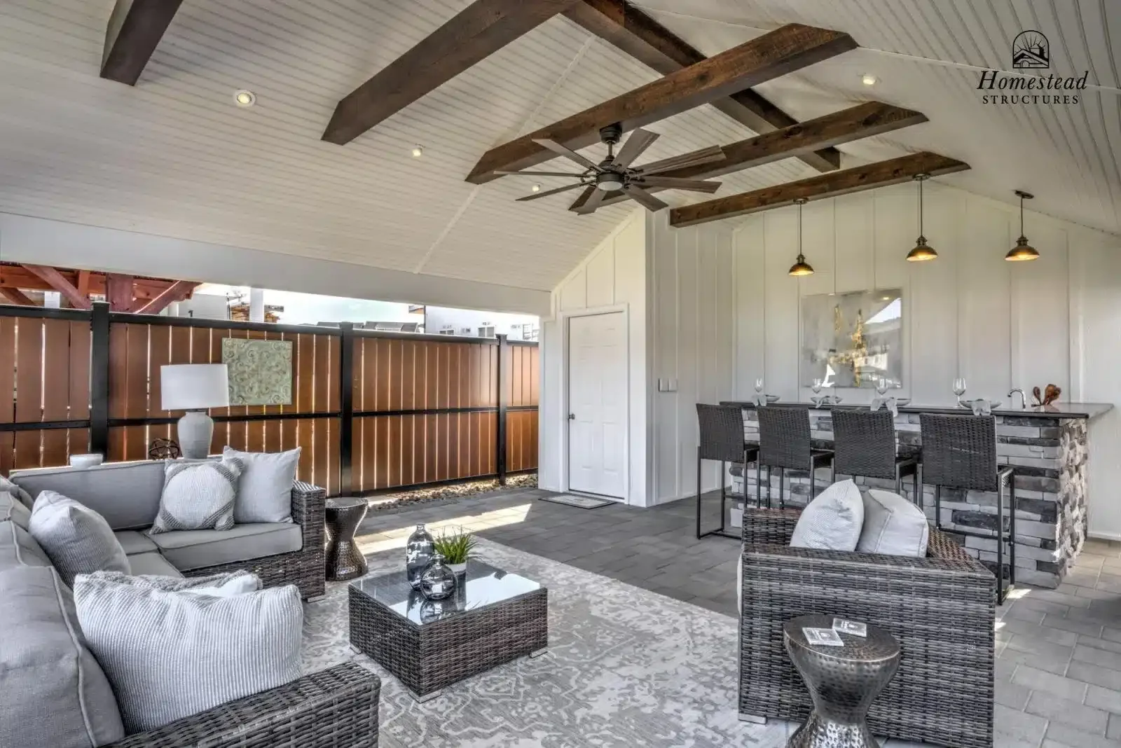 An outdoor living space with a vaulted ceiling featuring exposed wooden beams, a ceiling fan, and recessed lighting. There are two areas: a seating area with woven rattan furniture, cushioned chairs, a coffee table, and decorative vases; and a bar co