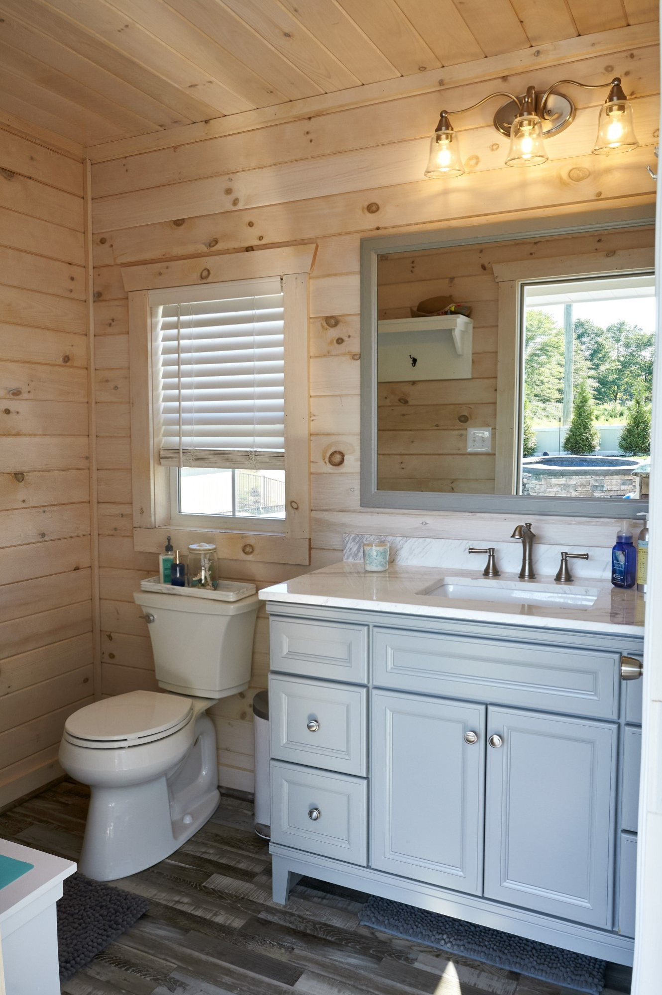 A rustic bathroom with wooden walls and ceiling, featuring a white toilet and a light blue vanity with a marble countertop, a mirror, and natural light streaming through a window.