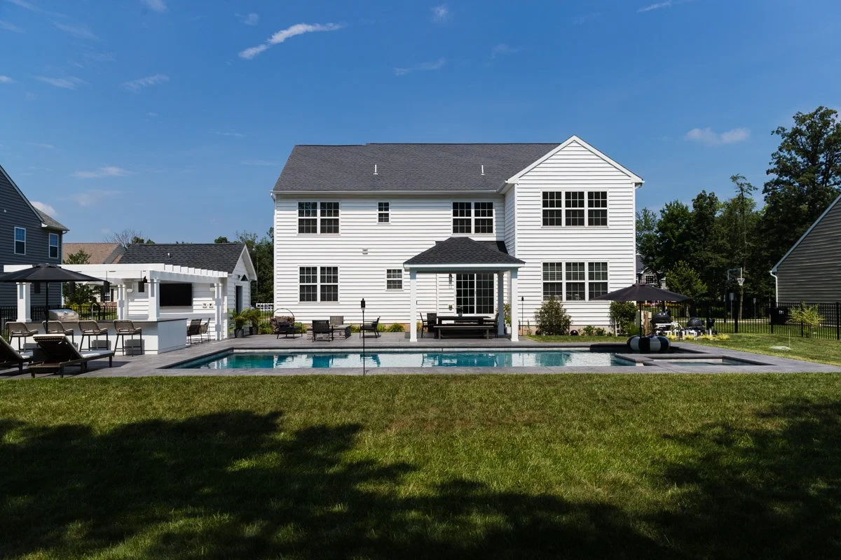 Backyard with swimming pool, patio furniture, and a white house with gray roof under a blue sky.
