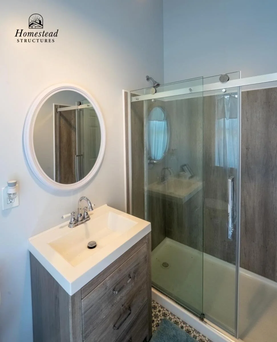 Bathroom with a small vanity, oval mirror, glass shower enclosure, wood-look shower walls, and a window with curtains.