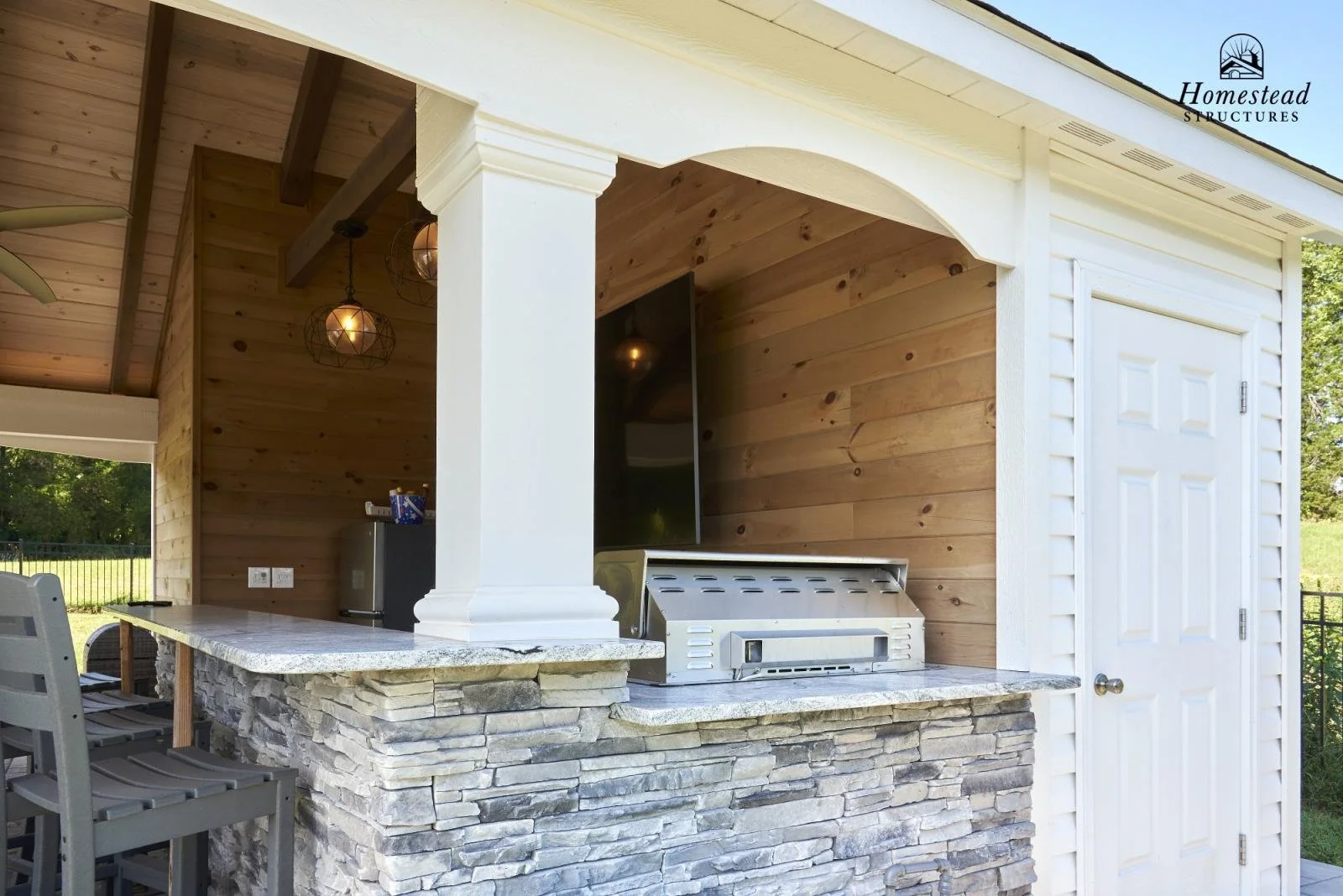 Outdoor kitchen with a stone counter, built-in grill, and decorative hanging lights, enclosed by a wooden wall with a white door, in a backyard setting.