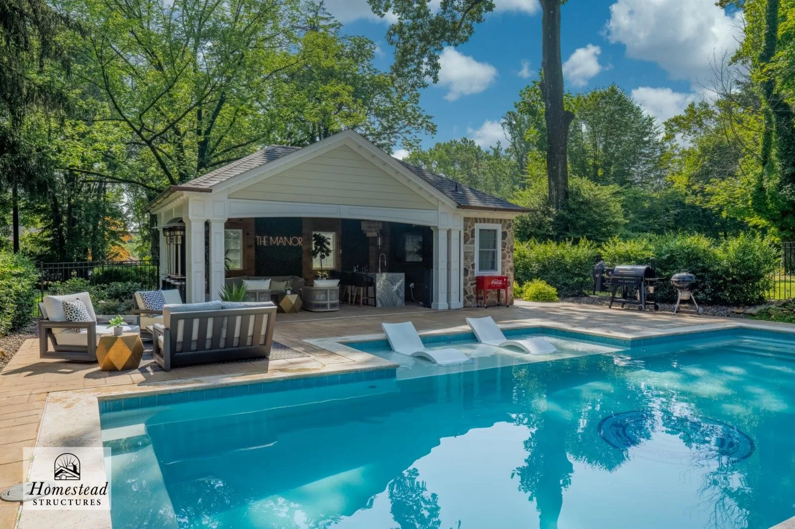 A backyard with a swimming pool, two built-in water slides, and an outdoor seating area with chairs and a small table under a pool house. The pool house has a stone and light yellow siding exterior, and there are trees and greenery surrounding the ar