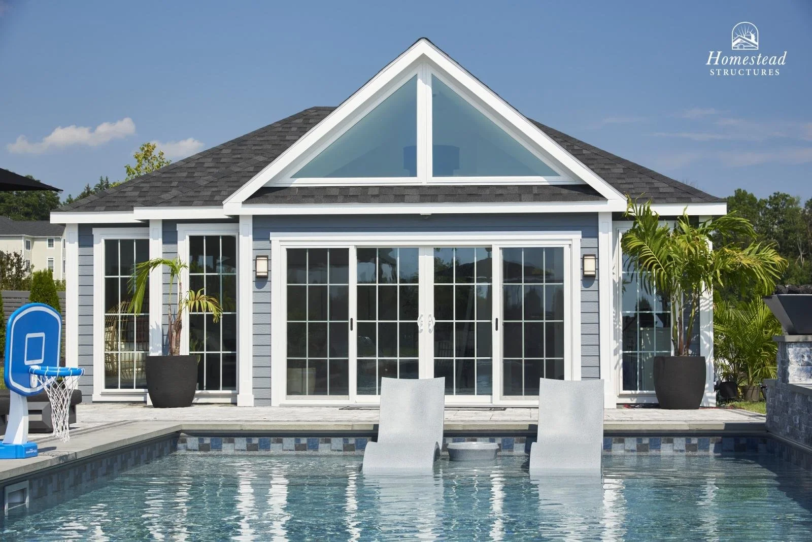 Backyard with a modern house featuring large glass sliding doors, a swimming pool with two lounge chairs partially submerged, potted palm trees, and a backyard basketball hoop, under a clear blue sky.